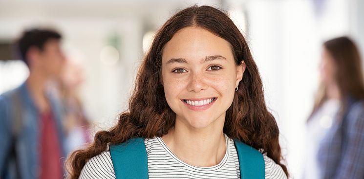 A Teenage Girl Smiling At The Camera While Wearing Her Backpack At School.